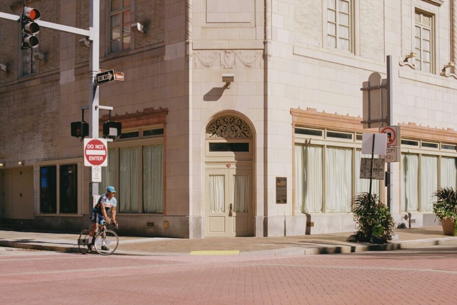 Um Homem Andando De Bicicleta Por Uma Rua Ao Lado De Um Predio Alto ihbJtM1vDho