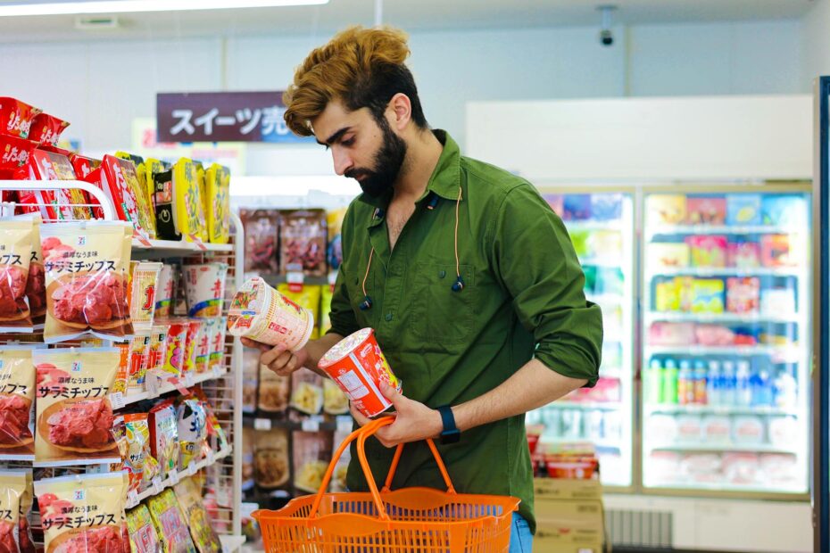 Adult man choosing cup noodles in a vibrant supermarket aisle holding an orange basket.