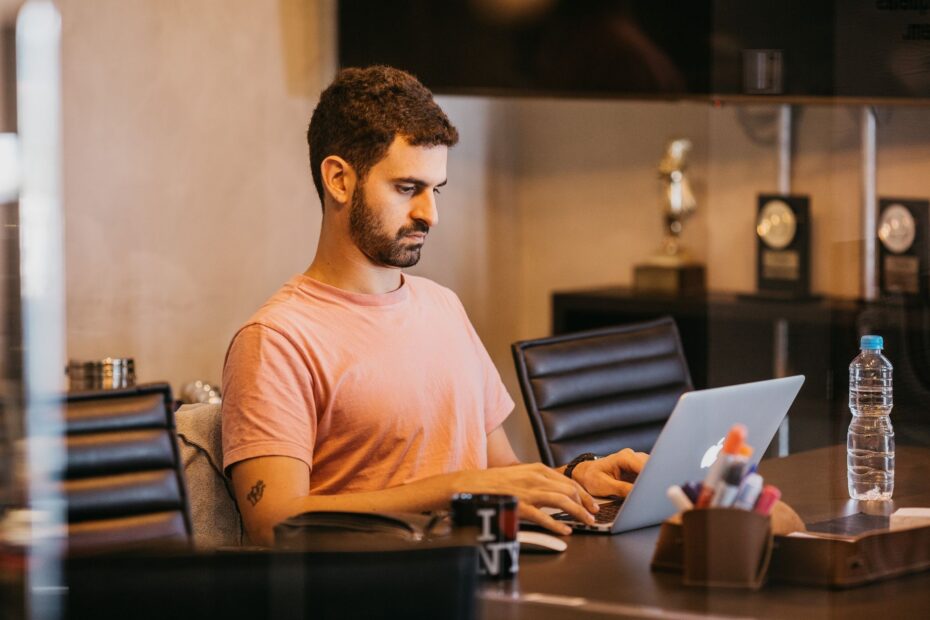 Man with short hair in pink t-shirt working on a laptop in a modern office setting.