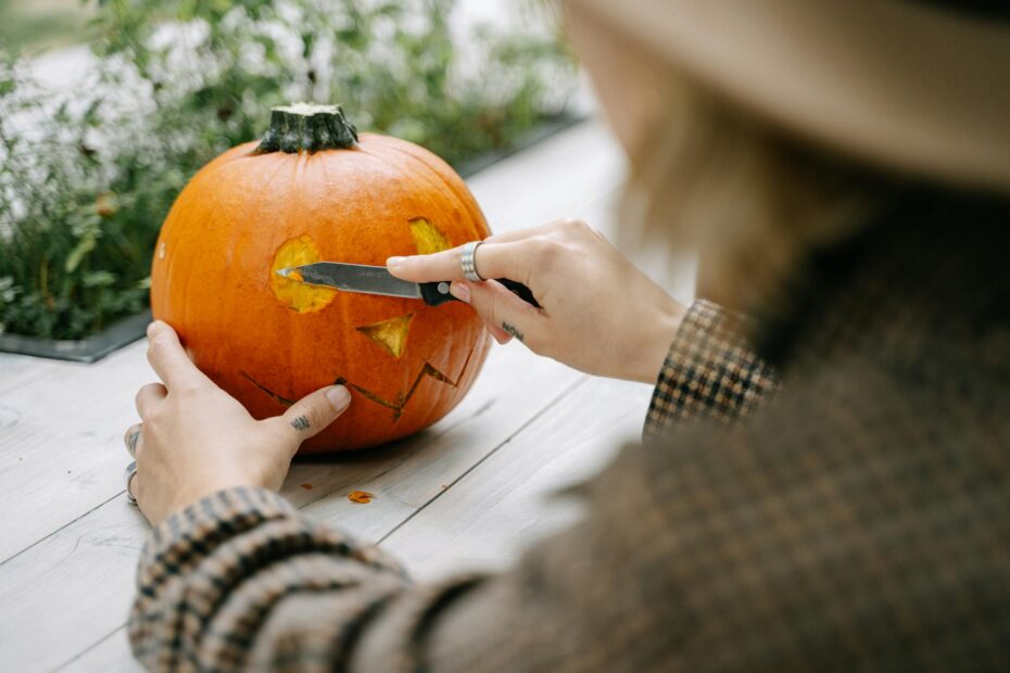 A woman carves a Halloween pumpkin with a knife on a wooden table outdoors.