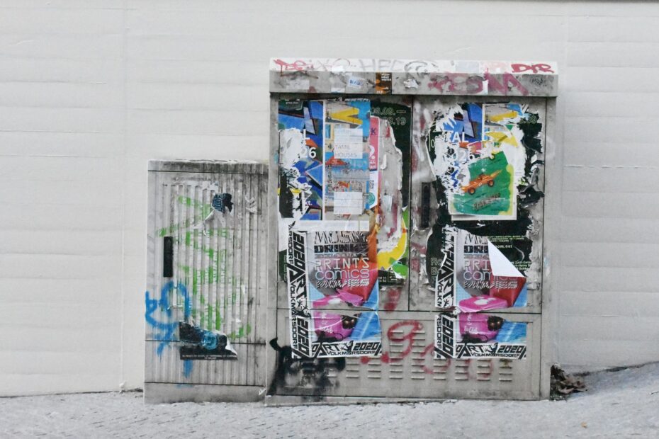 Graffitied and poster-covered electrical box in an urban setting, showcasing street art.