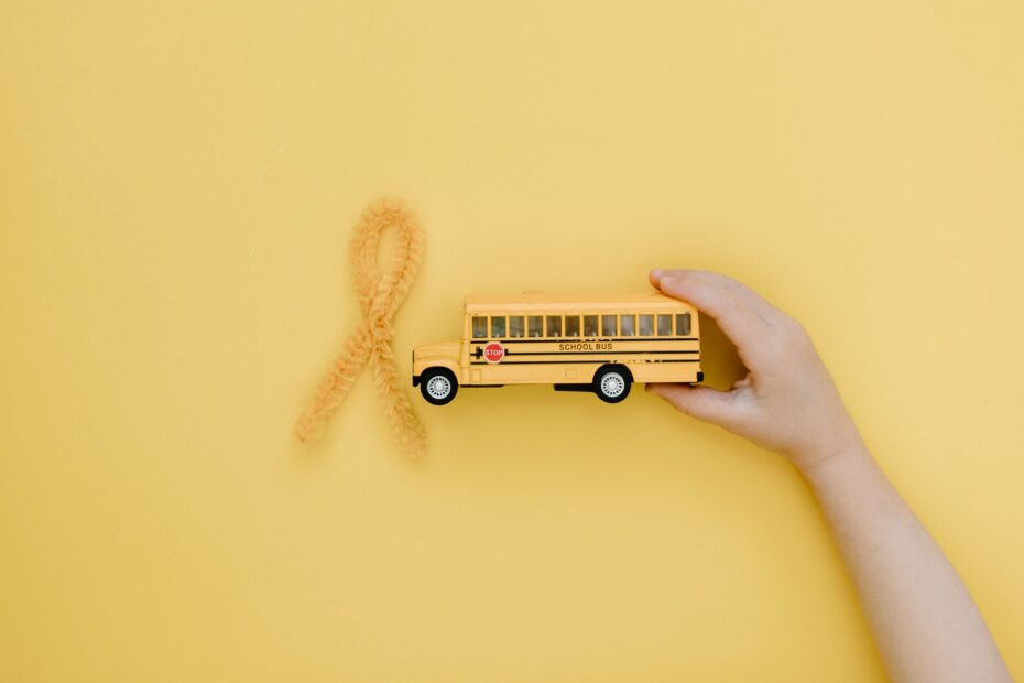 Hand holding toy school bus near yellow ribbon for cancer awareness on yellow background.