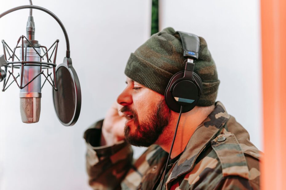 A bearded man wearing camo attire records music using a condenser microphone in a studio.
